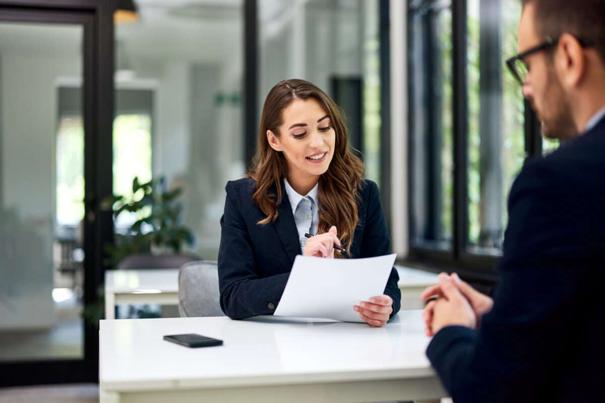 Businesswoman at a desk, looking happy and holding paper in front of job candidate